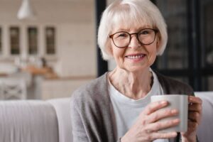 Elderly woman with glasses smiling while holding a cup of coffee in a cozy home setting.