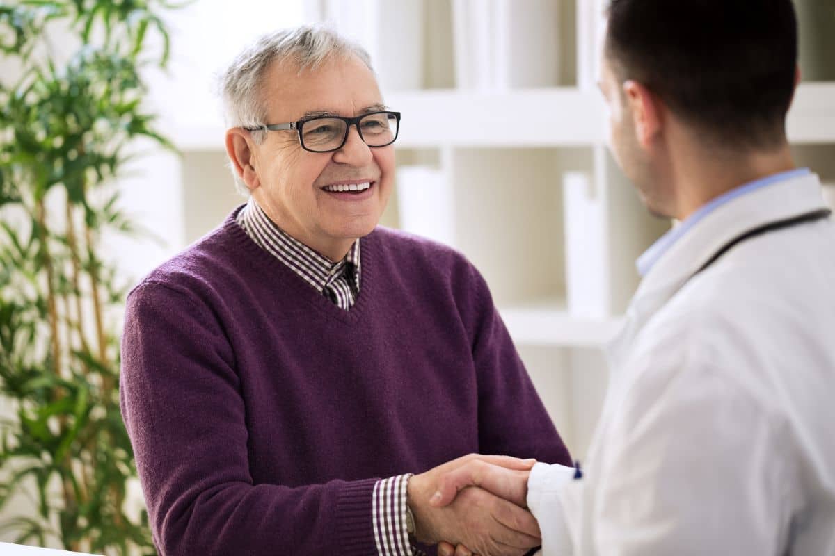 Elderly man in a purple sweater smiling while shaking hands with a doctor during a medical consultation.